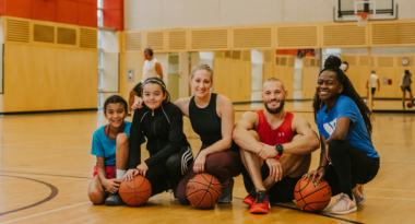 A group of people are sitting on the floor in a gym with basketballs.