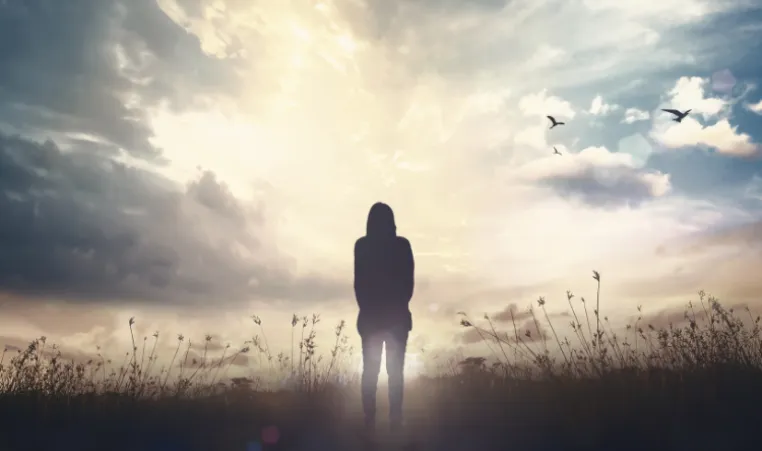 The silhouette of a person looking into the distance while standing in a field facing the sun