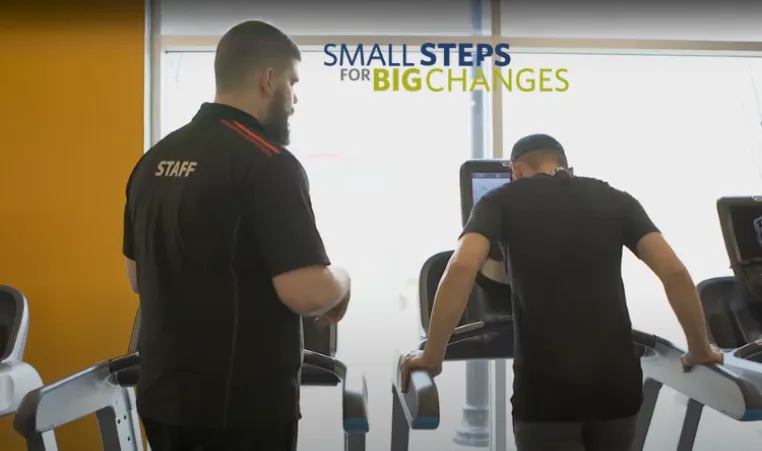 Two people are standing on treadmills with one person directing the other one. The Small Steps for Big Changes logo is above the second person.