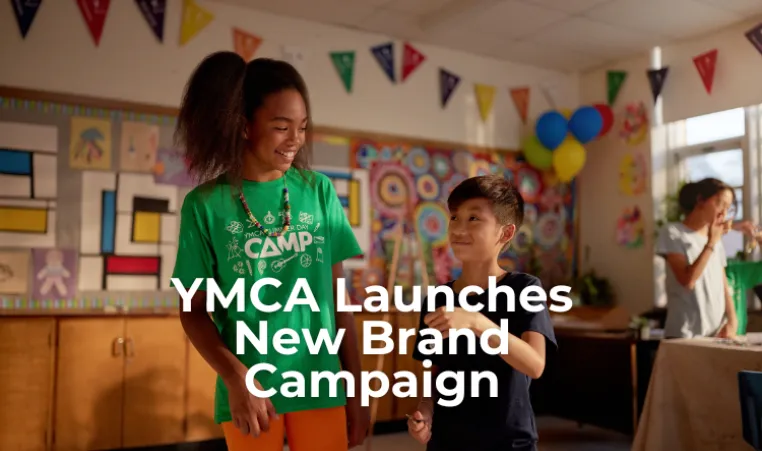Text: YMCA Launches New Brand Campaign. Image: A girl and a boy are standing inside a child care room smiling at each other.