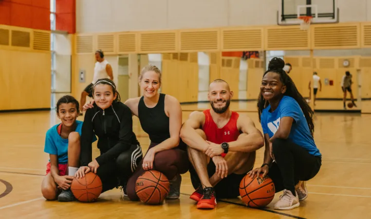 A group of people are sitting on the floor in a gym with basketballs.