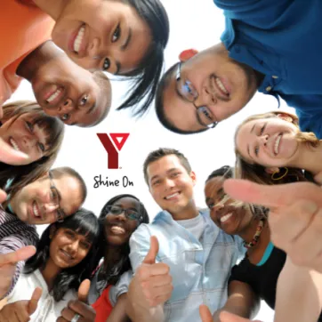 A group of young people are looking down toward the camera and are smiling and holding their thumbs up. The YMCA Shine On logo is in the middle