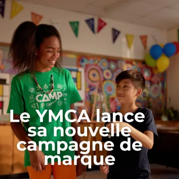 Texte : LE YMCA LANCE SA NOUVELLE CAMPAGNE DE MARQUE. Une fille et un garçon se tiennent debout dans une salle de garderie en souriant.