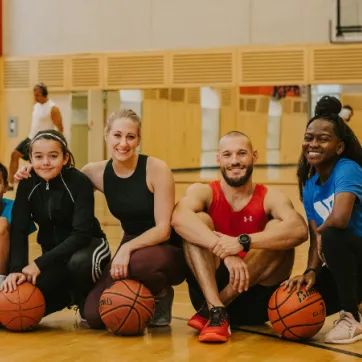 A group of people are sitting on the floor in a gym with basketballs.