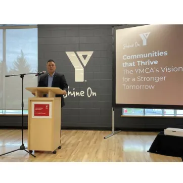 Peter Dinsdale is standing at a podium speaking. There is a screen projected behind him with the words:: Communities that Thrive. The YMCA's Vision for a Stronger Tomorrow
